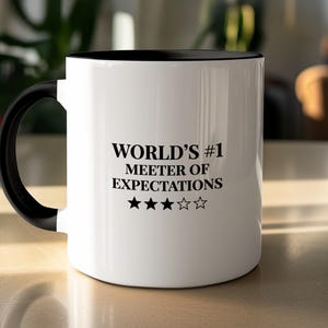 May include: A white ceramic mug with a black handle and interior. The front features black text that reads WORLD'S #1 MEETER OF EXPECTATIONS, followed by a row of five star icons, with the first three stars filled in black and two stars outlined.