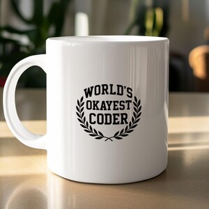Peut inclure: Une tasse en céramique blanche avec une anse noire. La tasse porte l'inscription "WORLD'S OKAYEST CODER" en noir, dans une police stylisée, entourée d'une couronne de laurier. La tasse est posée sur une surface claire.