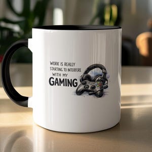 Può includere: Tazza in ceramica bianca con interno e manico neri. La tazza presenta la scritta "WORK IS REALLY STARTING TO INTERFERE WITH MY GAMING" insieme a un'illustrazione di un controller di gioco e cuffie.