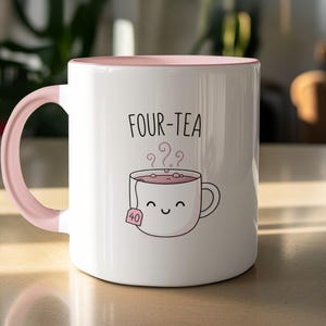 May include: White ceramic mug with a pink interior and handle. The mug features the text "FOUR-TEA" above a cartoon teacup with steam and a tea bag. The tea bag has the number "40" on it.