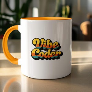 May include: White ceramic mug with an orange handle and rim. The mug features the text "Vibe Coder" in a retro font with yellow, orange, teal, and brown colours. The mug is sitting on a light-coloured surface.