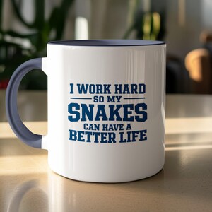 May include: White ceramic mug with a blue interior and handle. The mug features the text "I WORK HARD SO MY SNAKES CAN HAVE A BETTER LIFE" in blue block letters. The mug is sitting on a light-coloured surface.