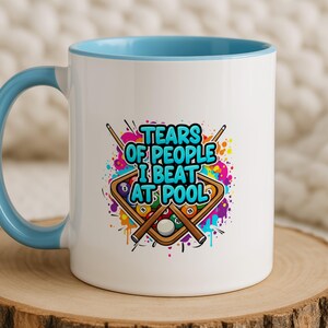 Può includere: Tazza in ceramica bianca con manico e bordo interno azzurro. La tazza presenta la scritta "TEARS OF PEOPLE I BEAT AT POOL" con una grafica colorata di palle e stecche da biliardo.