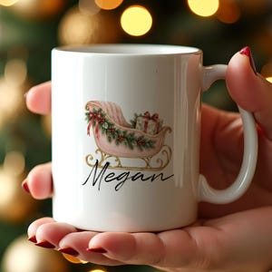 May include: A white ceramic mug featuring a printed illustration of a pink sleigh decorated with green pine branches, red berries, and a gift box. The name Megan is printed in black cursive script below the sleigh graphic on the mug surface.