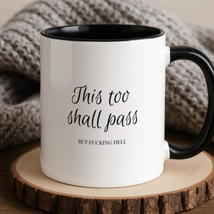 Pode incluir: Uma caneca de cerâmica branca com interior e alça pretos. A parte frontal da caneca apresenta o texto impresso em preto "This too shall pass" em letra cursiva, seguido pelo texto "BUT FUCKING HELL" numa fonte sem serifa menor.