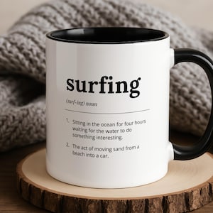 May include: White ceramic mug with a black interior and handle. The mug displays the word "surfing" in black, with a humorous definition below. The mug is on a wooden coaster, with a grey knitted blanket in the background.