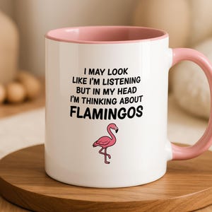 May include: A white ceramic mug with a pink interior and handle. The front features black text that reads I MAY LOOK LIKE I'M LISTENING BUT IN MY HEAD I'M THINKING ABOUT FLAMINGOS, accompanied by a small illustration of a pink flamingo below the text.
