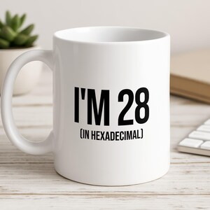 Könnte beinhalten: Ein weißer Keramikbecher mit dem schwarz aufgedruckten Text I'M 28 (IN HEXADECIMAL), der mittig auf der Vorderseite platziert ist. Der Becher hat eine glatte Oberfläche, einen geschwungenen Henkel auf der linken Seite und einen zylindrischen Körper.