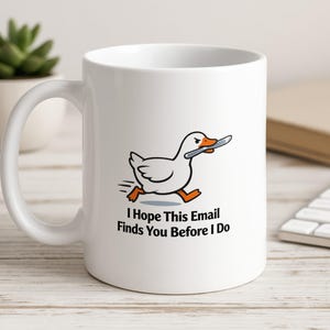 May include: White ceramic mug with a cartoon duck holding a knife, and the text "I Hope This Email Finds You Before I Do". The duck is running. A small succulent plant in a white pot is visible in the background. The mug is on a wooden surface.