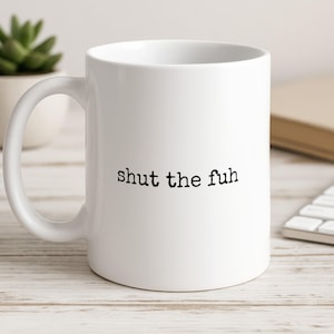 May include: A white ceramic mug with a curved handle featuring the black text "shut the fuh" printed in a typewriter-style font on the front centre. The mug has a smooth, glossy finish and a standard cylindrical shape with a wide, open top rim.