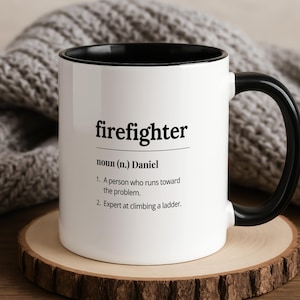 Peut inclure: Mug blanc avec intérieur et anse noirs, portant l'inscription "firefighter" et une définition. Le mug est posé sur un dessous de verre en bois, avec une couverture en tricot grise en arrière-plan.
