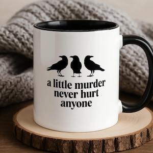 May include: White ceramic mug with a black interior and handle. The mug features three black raven silhouettes above the phrase "a little murder never hurt anyone" in a black, bold font. The mug sits on a wooden coaster.