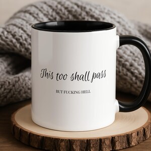 May include: White ceramic mug with a black interior and handle. The mug has the text "This too shall pass" above "BUT FUCKING HELL" in black script. The mug is on a wooden coaster.
