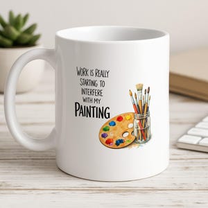 May include: White ceramic mug with the text "Work is really starting to interfere with my painting." The mug features a colourful artist's palette, paintbrushes, and a small jar. A succulent plant and keyboard are in the background.