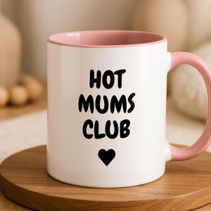 Op de afbeelding: Een witte keramische mok met een zachtroze binnenkant en handvat. De voorzijde van de mok bevat de zwarte tekst "HOT MUMS CLUB" in een vet, afgerond lettertype, geplaatst boven een klein, effen zwart harticoon gecentreerd bij de basis.