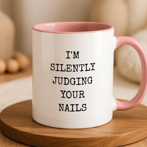Könnte beinhalten: Ein weißer Keramikbecher mit zartrosa Innenseite und passendem rosa Henkel. Auf der Vorderseite befindet sich ein schwarzer Text im Schreibmaschinenstil, der I'M SILENTLY JUDGING YOUR NAILS lautet und in vier zentrierten Zeilen gedruckt ist.
