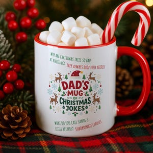 May include: A white ceramic mug with a red rim and handle, filled with white marshmallows and a red and white candy cane. The mug features green and red text reading DAD'S MUG OF CHRISTMAS JOKES, along with two jokes and festive holiday graphics.