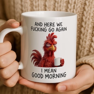 May include: A white ceramic mug featuring a graphic of a grumpy red rooster holding a small white mug. The text above the rooster reads AND HERE WE FUCKING GO AGAIN and the text below reads I MEAN GOOD MORNING in black capital letters.