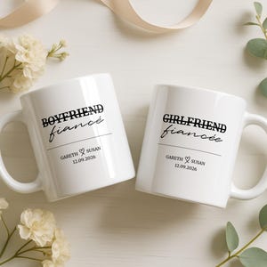 Könnte beinhalten: Zwei weiße Keramiktassen mit schwarzem Text. Eine Tasse trägt die Aufschrift BOYFRIEND fiancé mit GARETH ♡ SUSAN 12.09.2026. Die andere Tasse trägt die Aufschrift GIRLFRIEND fiancée mit GARETH ♡ SUSAN 12.09.2026. Beide Tassen haben einen Henkel.