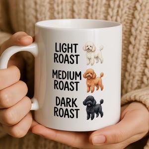 Peut inclure: Un mug en céramique blanche orné de trois illustrations de caniches accompagnées de texte. Le caniche du haut est crème à côté de LIGHT ROAST, celui du milieu est marron à côté de MEDIUM ROAST, et celui du bas est noir à côté de DARK ROAST.