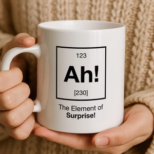 May include: A white ceramic mug featuring a black periodic table-style graphic with the number 123 at the top, the letters Ah! in the centre, the number 230 in brackets below, and the text The Element of Surprise! printed at the bottom.