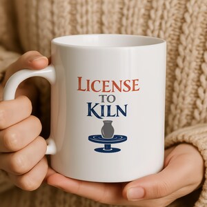 Może przedstawiać: Biały ceramiczny kubek z napisem "License to Kiln" w kolorze czerwonym i niebieskim. Kubek przedstawia grafikę wazonu ceramicznego na kole garncarskim. Kubek ma duży, zakrzywiony uchwyt.