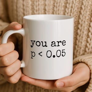 May include: White ceramic mug with the text "you are p < 0.05" in black. The mug has a curved handle and is held by a person wearing a beige jumper. The mug is a standard size for coffee or tea.