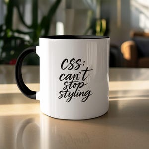 Puede incluir: Taza de cerámica blanca con interior y asa negros. La taza presenta el texto "CSS: can't stop styling" en negro, con una fuente manuscrita. La taza está sobre una superficie clara.
