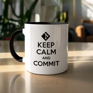 Może przedstawiać: Biały ceramiczny kubek z czarnym wnętrzem i uchwytem. Kubek zawiera czarny napis "KEEP CALM AND COMMIT" oraz czarno-białą grafikę. Kubek stoi na jasnej powierzchni.