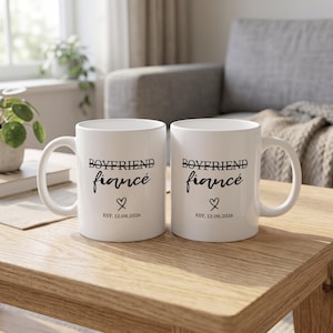 Two white mugs with "Boyfriend" crossed out and "fiancé" printed on them with a date.