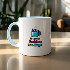 May include: White ceramic coffee mug with a light blue handle and rim. The mug features a pixelated design of a coffee cup and the text "I turn Coffee into Bugs" in a colorful, pixelated font. The mug is on a light-coloured surface.
