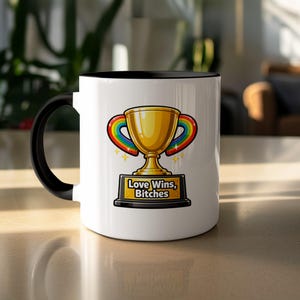 Może przedstawiać: Biały ceramiczny kubek z czarnym wnętrzem i uchwytem. Kubek ma złoty wzór trofeum z tęczowym łukiem i napisem "Love Wins, Bitches" na czarnej podstawie. Projekt zawiera złote gwiazdy.