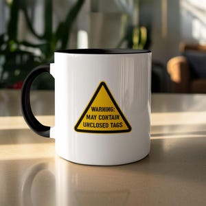 Könnte beinhalten: Weißer Keramikbecher mit schwarzem Innenraum und Henkel. Der Becher hat ein gelbes Warnschild mit schwarzem Text: "WARNING: MAY CONTAIN UNCLOSED TAGS."