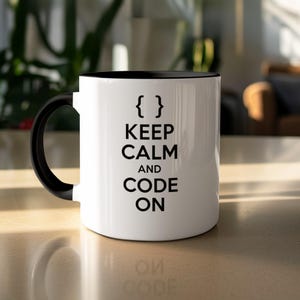 Peut inclure: Mug en céramique blanche avec intérieur et anse noirs. Le mug porte l'inscription "KEEP CALM AND CODE ON" en noir, avec des accolades au-dessus. Le mug est posé sur une surface claire.