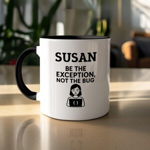 May include: White ceramic mug with a black interior and handle. The mug features the name "SUSAN" in bold black letters, along with the phrase "BE THE EXCEPTION, NOT THE BUG" and a graphic of a person holding a laptop.