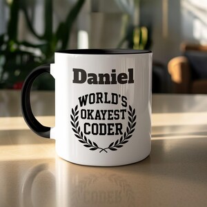 Peut inclure: Mug en céramique blanche avec intérieur et anse noirs. Le mug affiche le nom "Daniel" au-dessus du texte "WORLD'S OKAYEST CODER" à l'intérieur d'une couronne de laurier.