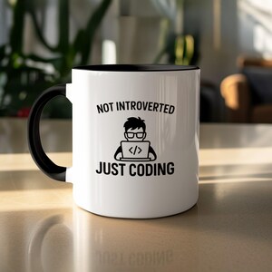 May include: White ceramic mug with a black interior and handle. The mug features the text "NOT INTROVERTED JUST CODING" above an illustration of a person coding on a laptop. The mug is sitting on a light-colored surface.