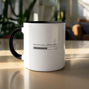 May include: White coffee mug with a black interior and handle. The mug has the text "installing coffee.exe" and a black progress bar graphic. The mug is on a light-coloured surface.