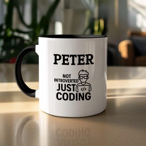 Puede incluir: Taza de cerámica blanca con interior y asa negros. La taza presenta el nombre "PETER" en letras negras en negrita, junto con el texto "NOT INTROVERTED JUST CODING" y un gráfico de una persona con gafas y símbolos de codificación.