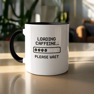 May include: White coffee mug with a black interior and handle. The mug has the text "LOADING CAFFEINE..." above a loading bar graphic and the words "PLEASE WAIT" in a pixelated font.