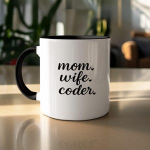 Peut inclure: Mug en céramique blanche avec intérieur et anse noirs. Le mug porte les mots "mom. wife. coder." en noir, dans une police manuscrite. Le mug est posé sur une surface claire.