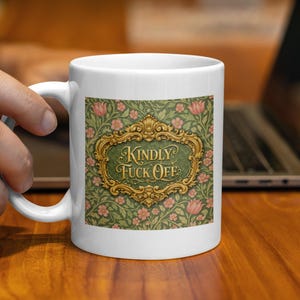 Può includere: Tazza in ceramica bianca con un design decorativo. La tazza presenta uno sfondo verde con fiori rosa e un testo incorniciato in oro che recita "KINDLY FUCK OFF". La tazza è tenuta da una mano.