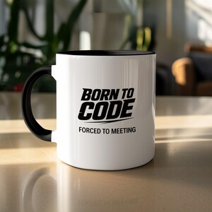 May include: White ceramic mug with a black interior and handle. The mug features the text "BORN TO CODE" above "FORCED TO MEETING" in a bold, black font. The mug is on a light-coloured surface.