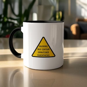 Może przedstawiać: Biały ceramiczny kubek z czarnym wnętrzem i uchwytem. Kubek posiada żółty znak ostrzegawczy z napisem: "WARNING: MAY CONTAIN UNMATCHED PARENTHESIS". Kubek stoi na jasnej powierzchni.