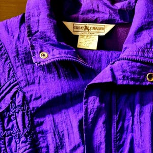 May include: A vibrant purple jacket with a high collar and a zipper closure. The jacket features gold-colored snaps and a label that reads "GREAT CAVALIER". The fabric has a textured appearance, and the design includes gathered fabric details.