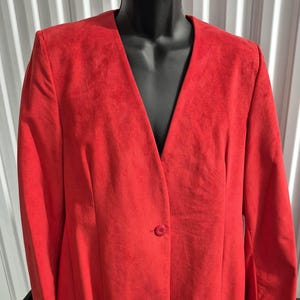 Pode incluir: Um blazer vermelho brilhante com decote em V e fecho de um botão. O casaco é exibido em um manequim preto e tem um design clássico e sob medida. O tecido parece ser um material macio semelhante a camurça.