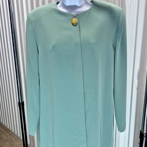 May include: A long-sleeved, mint green dress coat with a single gold button closure. The coat is displayed on a white mannequin and has a simple, elegant design. The fabric appears to be a smooth, lightweight material.