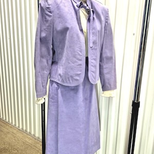 Puede incluir: Un traje de falda vintage de ante color lavanda. El traje incluye una chaqueta con cuello redondo y una falda a juego hasta la rodilla. La chaqueta se lleva sobre una blusa color crema con lazo. El traje se exhibe en un perchero.