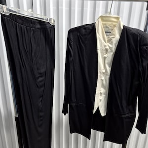 May include: A black two-piece suit hanging on a metal rack. The suit includes a long-sleeved jacket with a cream-colored blouse underneath, and matching black pants. The blouse has decorative buttons. The suit is made of a smooth, possibly satin-like material.