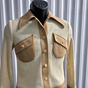 Cream-colored ribbed knit shirt with tan accents. The shirt features a wide tan collar, two front pockets, and tan trim along the button closure and cuffs. The shirt is buttoned up and displayed on a mannequin. Size 8, By Act III, a vintage company of Jonathan Logan.